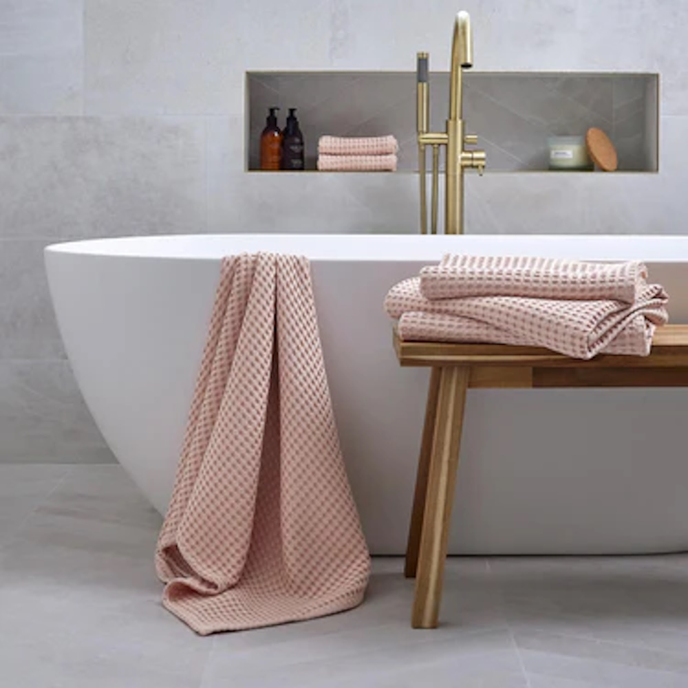 Pink waffle towels from Christy draped over the edge of a bath with some more folded on a wooden bench next to the bath