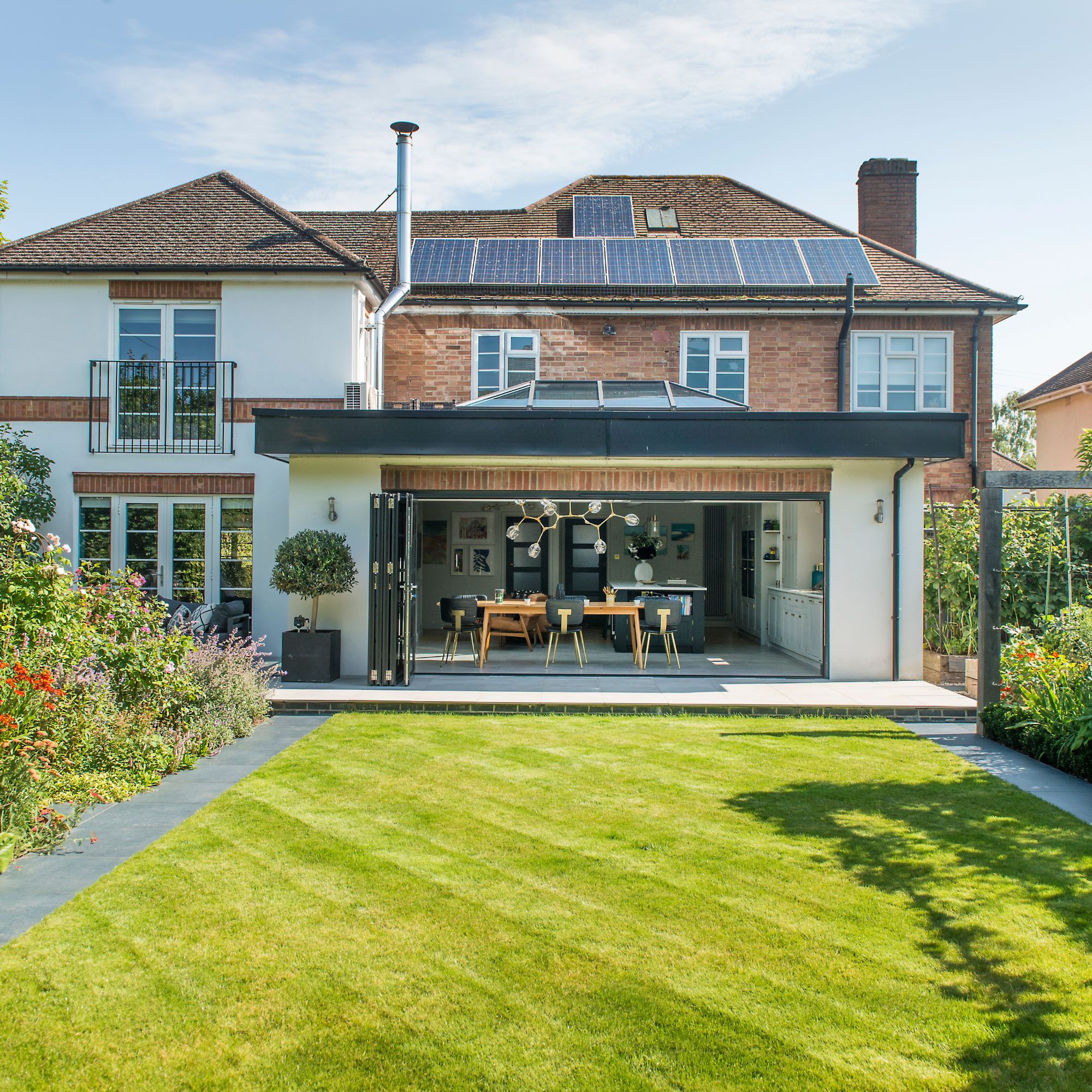 Rear elevation of large home with solar panels on the roof, a roof lantern on a flat roof extension, green lawn and plush garden