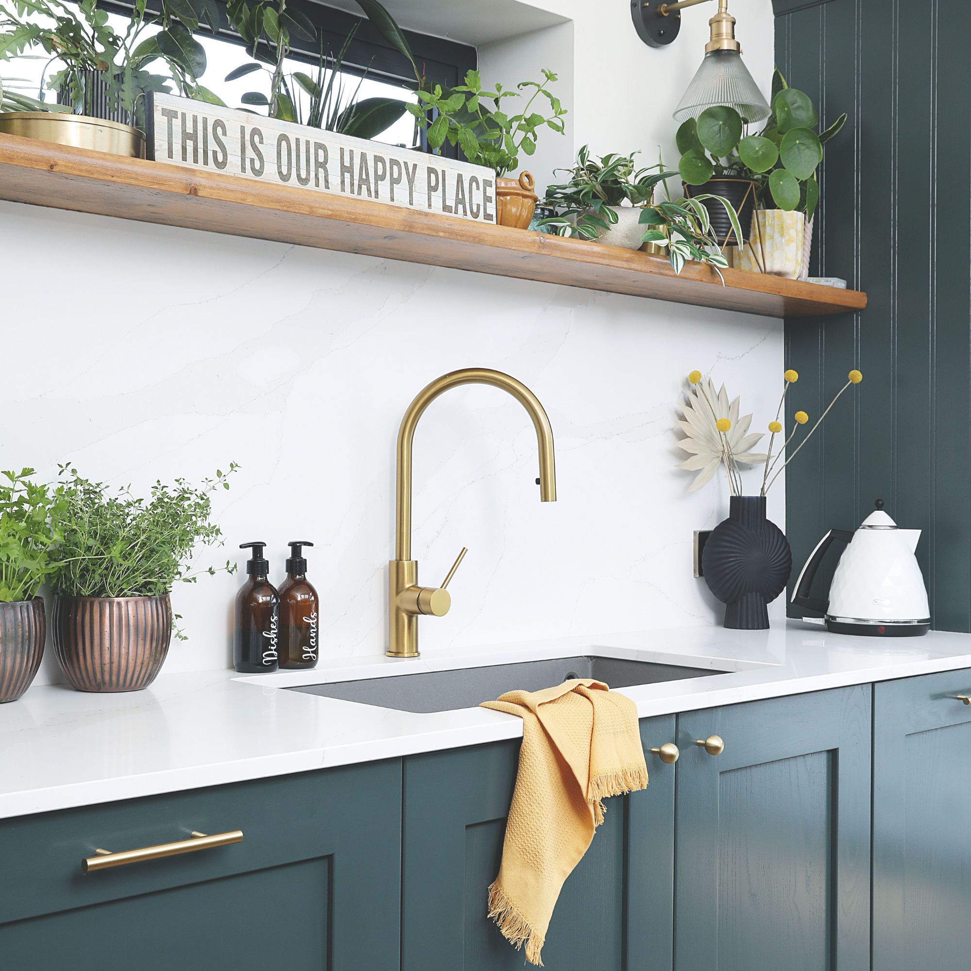 Gold kictchen tap, white countertops and navy blue cabienets. There is an ochre tea towel and lots of house plants.